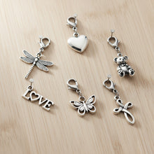 Set of silver cremation charms including a dragonfly, heart, teddy bear, butterfly, 'LOVE' word, and a cross on a wooden surface.